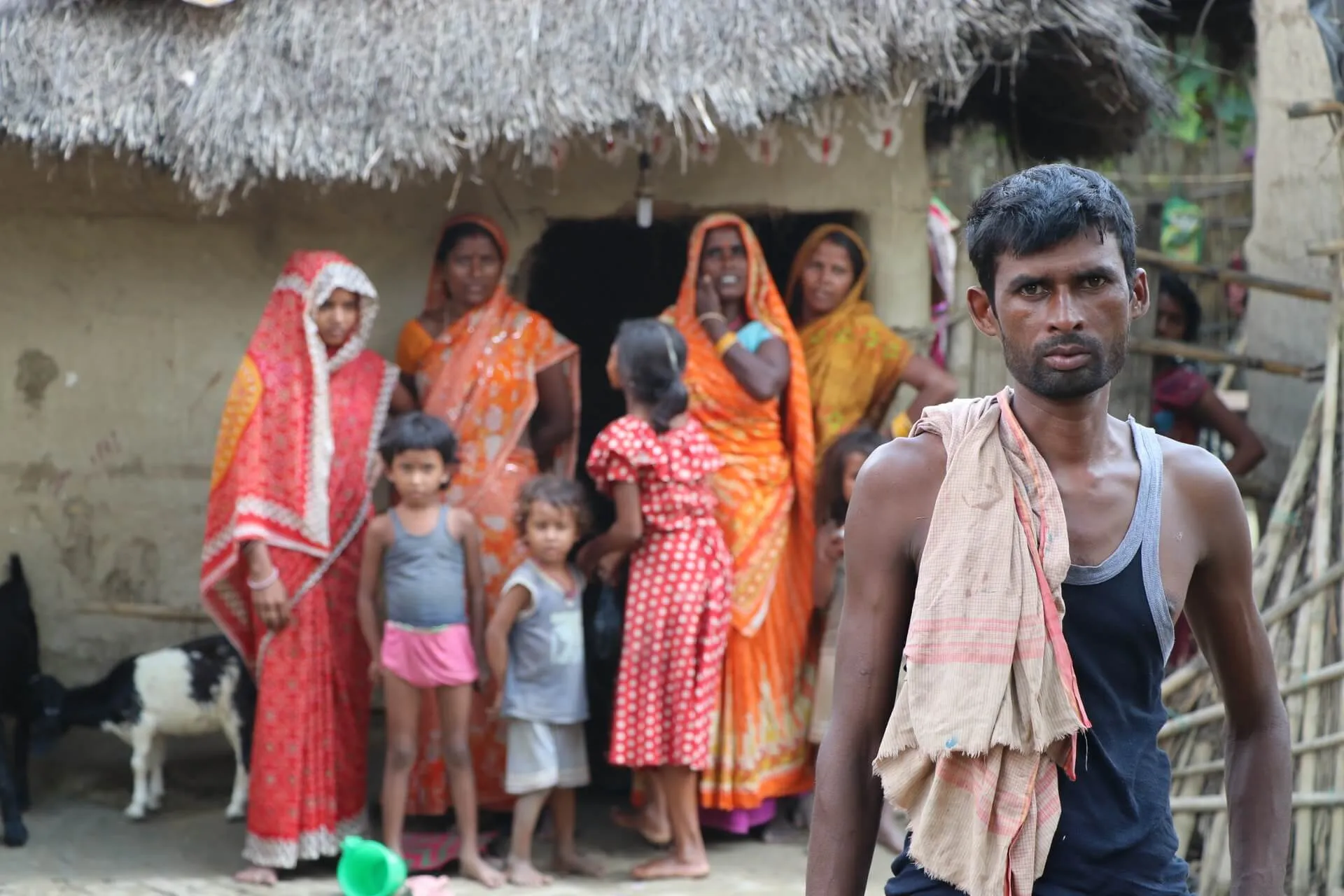 A family outside a thatched house in a village, a man standing in the foreground with a cloth over his shoulder.