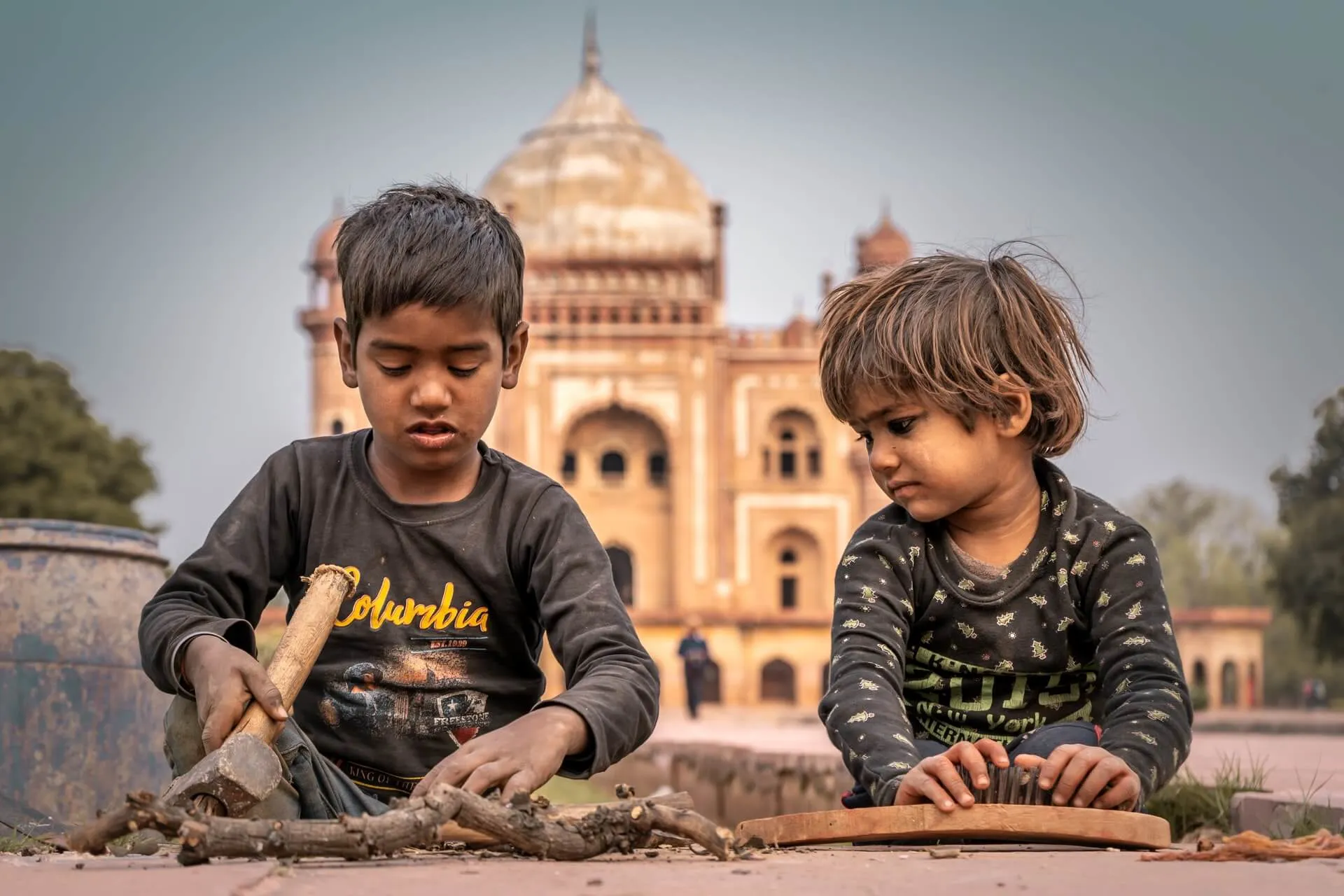 Two young children sitting on the ground in Delhi, with Safdarjung's Tomb behind them.