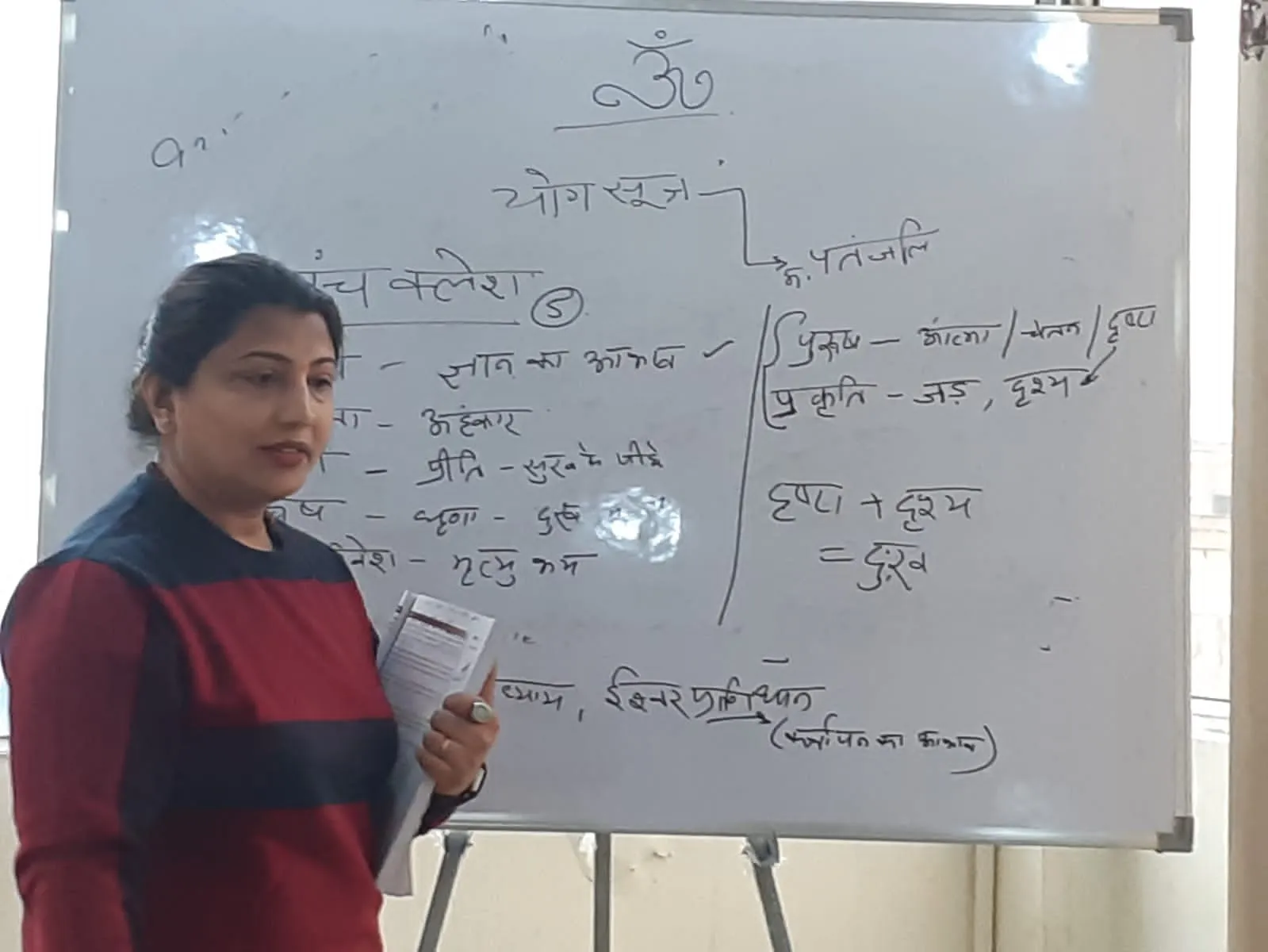 A TPNF teacher leading a Yoga Sutra class beside a whiteboard of handwritten Hindi notes.