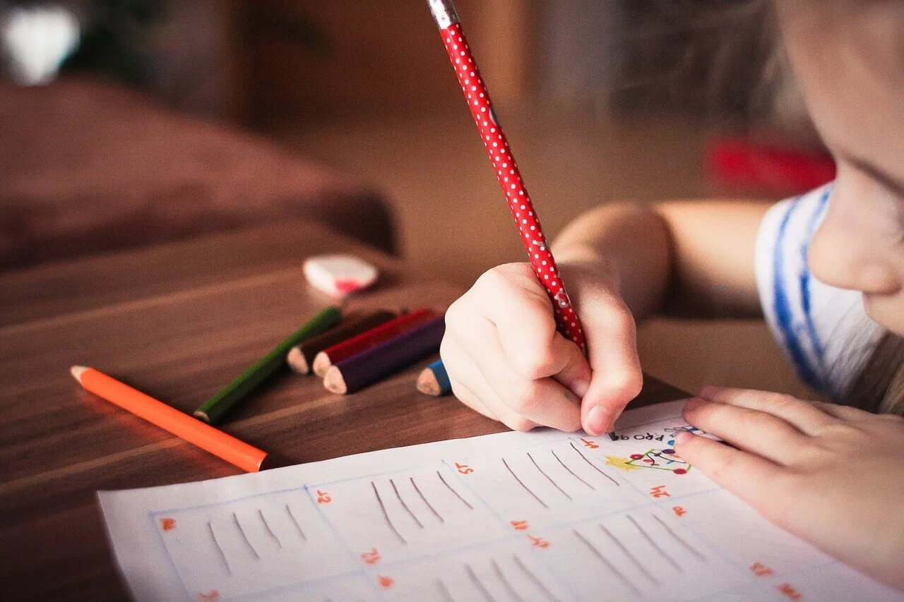A child writing in an exercise book with coloured pencils on the desk.